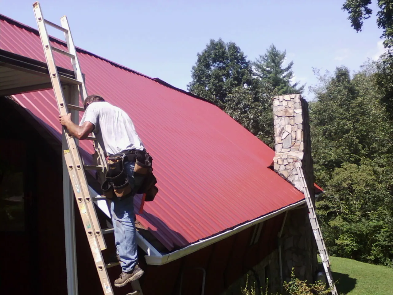 Roofer on a ladder beside a red metal roof and stone chimney — Soffit & Fascia Repair by Roofing Brothers in Bean Station